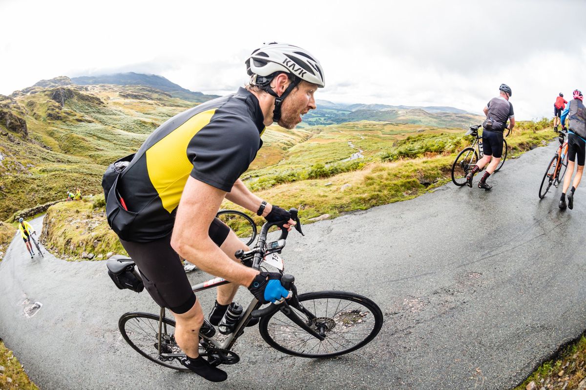Riders climbing a wet single-track pass in the Lake District during the Fred Whitton Challenge,
                  fells and low cloud behind.