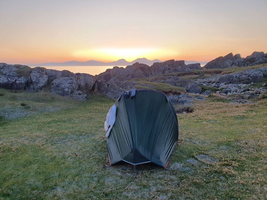 A small tent pitched on grass above a rocky shore, sun setting over the sea beyond.