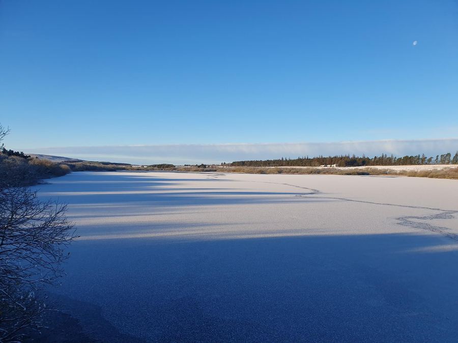A frozen reservoir with a pale ice sheet stretching to a low far shore under deep blue sky.