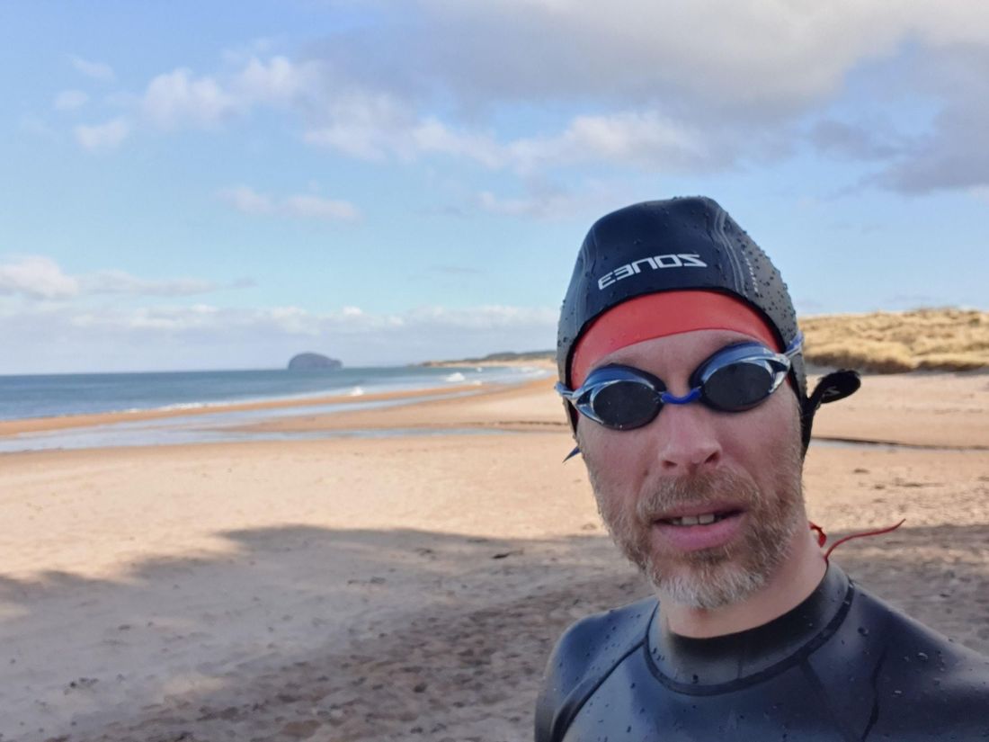 Gabriel Scarlett in a wetsuit and swimming goggles on a broad sandy beach, sea and an
                    island visible behind, under a bright winter sky.
