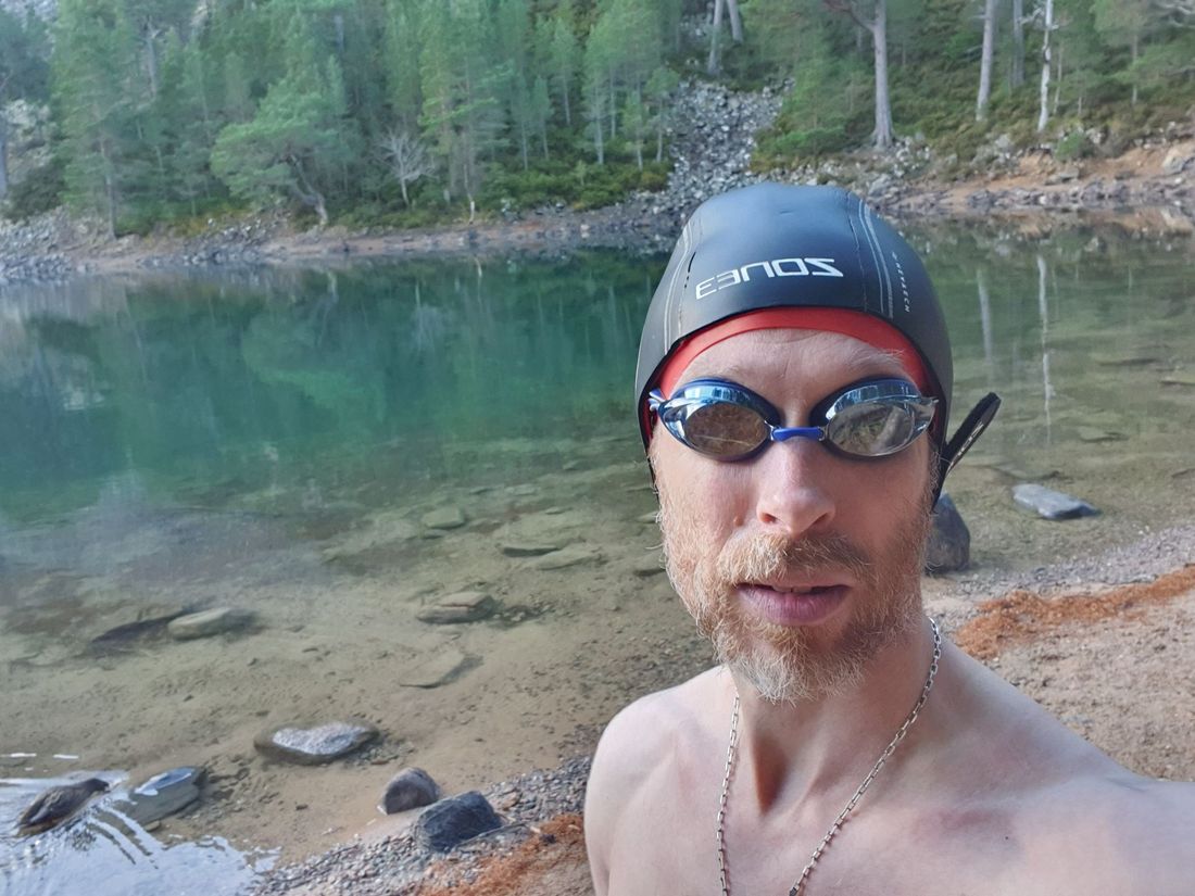 Gabriel Scarlett in a swim cap and goggles at the shore of An Lochan Uaine, the
                    green water and pine-covered slopes of the Cairngorms behind him.